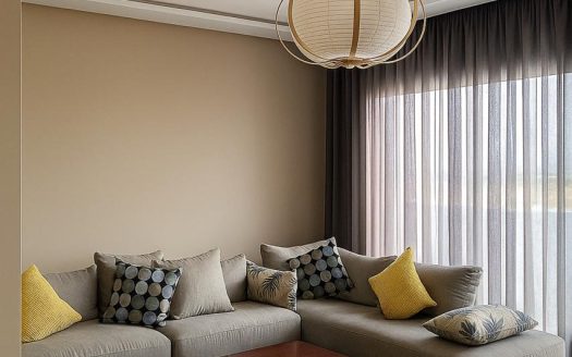 Salon lumineux du studio meublé au CIL Casablanca avec canapé d'angle gris, pouf vert olive et luminaire globe.